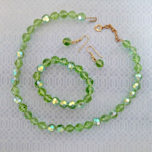 K&ouml;nnte beinhalten: Ein Set aus gr&uuml;nem, irisierendem Glasperlen-Schmuck, bestehend aus einer Halskette, einem Armband und Ohrringen. Die Halskette und das Armband bestehen aus facettierten Perlen, w&auml;hrend die Ohrringe einfache Tropfenformen haben.