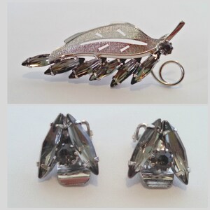 May include: A silver leaf-shaped brooch with a row of dark gray rhinestones. Matching earrings with three dark gray rhinestones each.