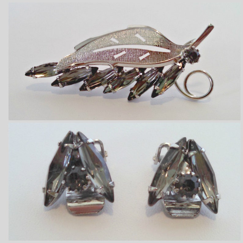 May include: A silver leaf-shaped brooch with a row of dark gray rhinestones. Matching earrings with three dark gray rhinestones each.