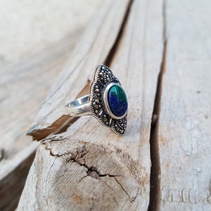 May include: Silver ring with an oval-shaped, two-tone blue and green stone. The ring has a decorative, ornate setting with small, round accents. The ring is set against a weathered wood background.