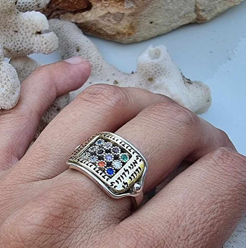 May include: A silver ring with a rectangular design featuring a gold-toned border and a cluster of colorful gemstones. The ring is inscribed with Hebrew text.