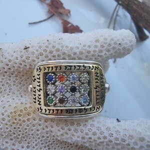 May include: A silver and gold ring with a square design featuring a variety of colorful gemstones and Hebrew lettering around the perimeter.