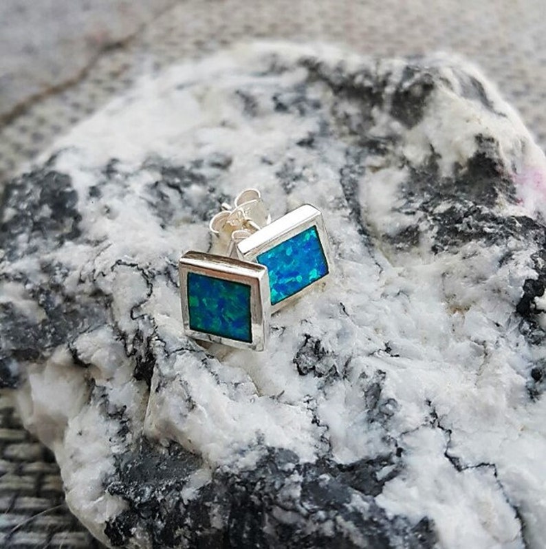May include: Square stud earrings with a vibrant blue and green opal inlay set in a silver frame. The earrings are displayed on a textured, gray and white stone surface, showcasing their color and design.