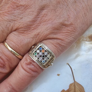 May include: A silver ring with a square-shaped design featuring a central cluster of colorful gemstones surrounded by smaller white gemstones. The ring is inscribed with Hebrew text.