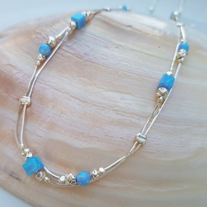 May include: A delicate silver chain necklace with two strands and blue opal beads. The necklace features both round and square shaped beads.