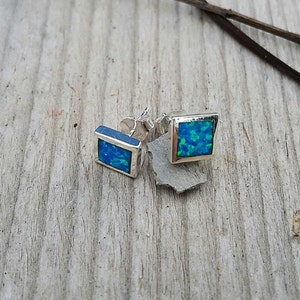 May include: A pair of square stud earrings with a silver frame. Each earring features a vibrant blue-green opal stone. The earrings are displayed on a weathered wooden surface, showcasing their design.