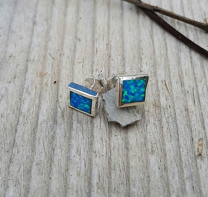 May include: A pair of square stud earrings with a silver frame. Each earring features a vibrant blue-green opal stone. The earrings are displayed on a weathered wooden surface, showcasing their design.