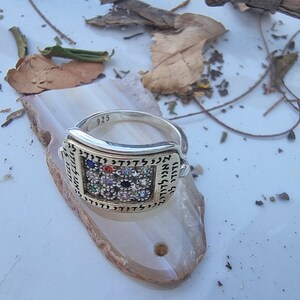 May include: A silver ring with a square-shaped design featuring a variety of colorful gemstones and Hebrew lettering. The ring is marked with "925" on the band.