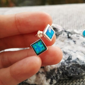 May include: A pair of silver stud earrings with a square, diamond-shaped design. Each earring features a vibrant blue opal stone set within a silver frame. The opal has a mix of blue and green hues, creating a shimmering effect.