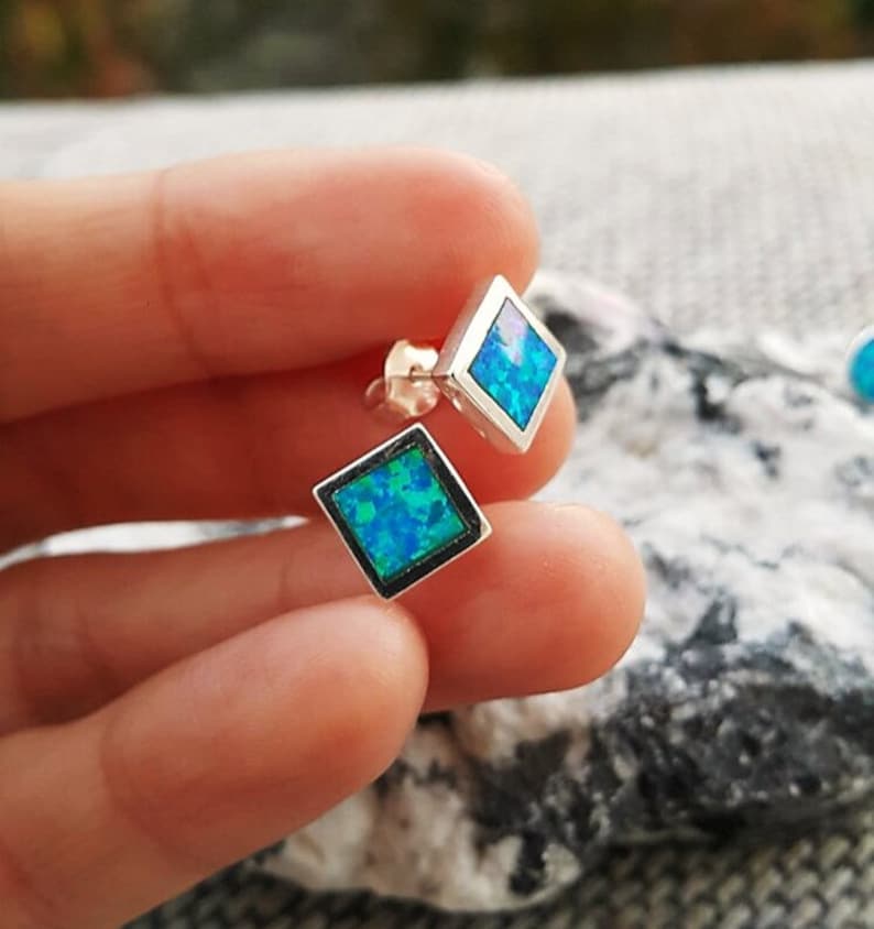May include: A pair of silver stud earrings with a square, diamond-shaped design. Each earring features a vibrant blue opal stone set within a silver frame. The opal has a mix of blue and green hues, creating a shimmering effect.