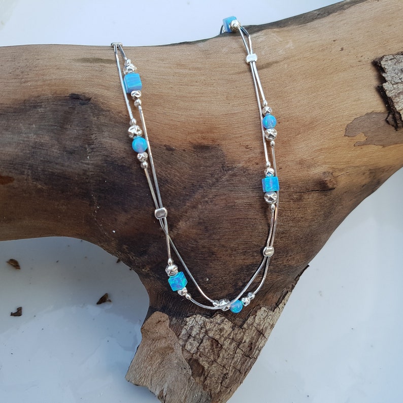 May include: A silver anklet with two delicate chains and blue opal beads. The anklet is displayed on a wooden surface.