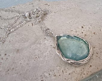 May include: A silver necklace featuring a teardrop-shaped pendant with a light blue stone. The stone is encased in a braided silver frame, and the chain is a delicate silver link. The necklace is displayed on a textured, light-colored surface.