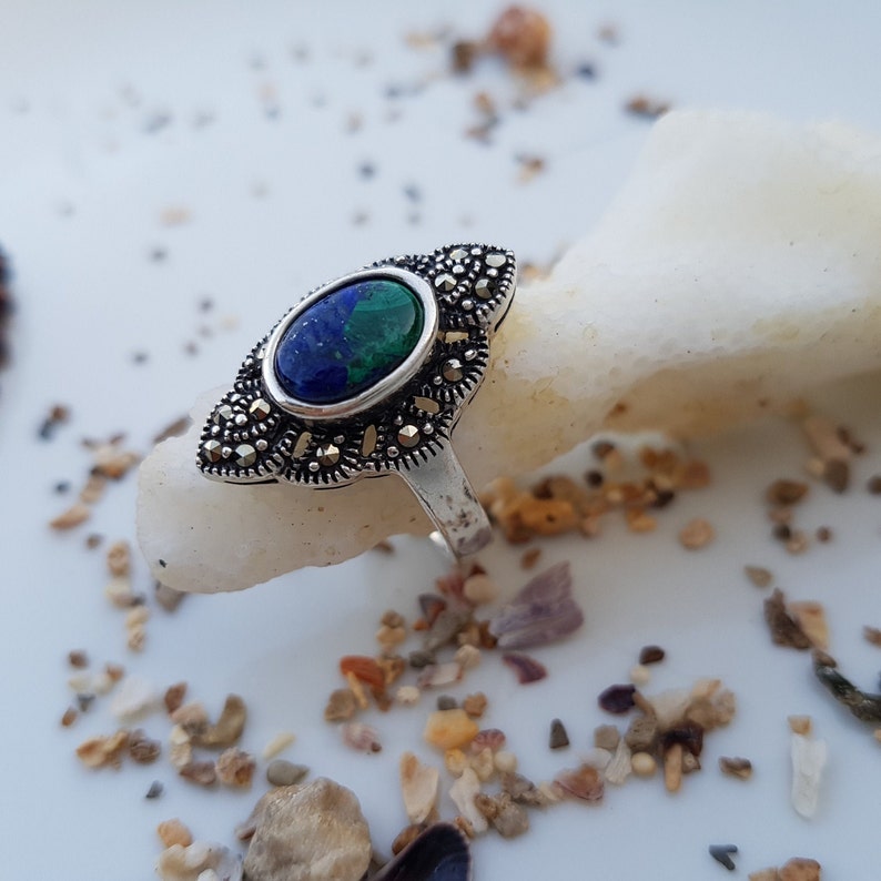 May include: A silver ring with a marcasite setting and an oval-shaped stone with blue and green hues. The ring is set on a white surface with small pebbles scattered around it.