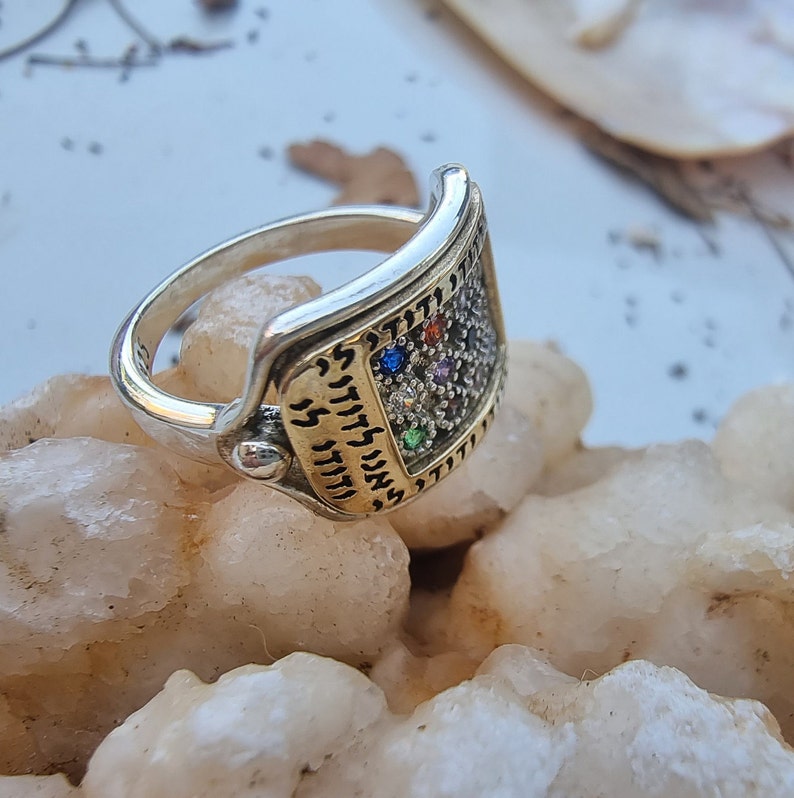 May include: A silver and gold ring with a rectangular design featuring Hebrew text and colorful gemstones. The ring is set on a white background.