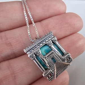 May include: A silver pendant necklace featuring a detailed architectural design with turquoise stone accents. The pendant showcases a square structure with columns, steps, and a central turquoise stone, suspended from a delicate chain.