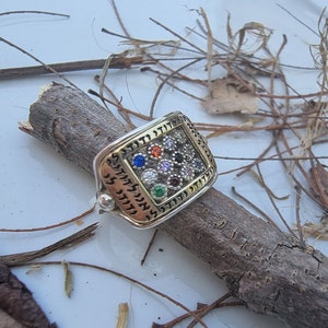 May include: A silver ring with a gold-toned square design featuring a Hebrew inscription and a cluster of colorful gemstones.