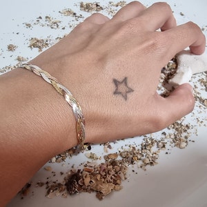 Puede incluir: Una pulsera trenzada de plata, oro y oro rosa en la muñeca de una persona. La pulsera está hecha de tres hebras de metal que están entrelazadas.