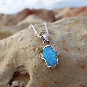 May include: A silver necklace featuring a Hamsa hand pendant with a vibrant blue opal inlay. The pendant is set against a textured, light brown stone, with the chain visible. The opal has shades of blue and green.
