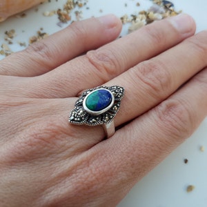 May include: A silver ring featuring an oval-shaped, blue and green gemstone. The gemstone is set in a decorative, marcasite-studded frame. The ring is displayed on a hand.
