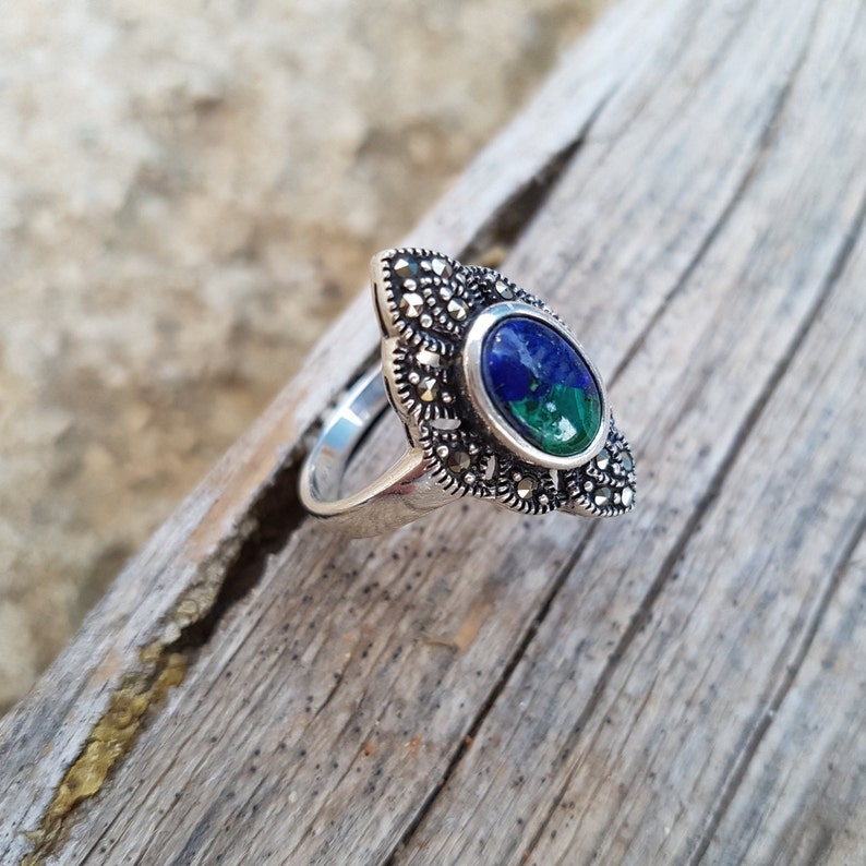 May include: A silver ring with an oval-shaped, two-tone blue and green gemstone. The ring is surrounded by small, round, silver-colored accents. The ring is set on a piece of weathered wood.