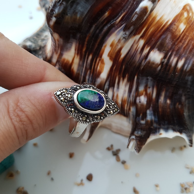 May include: A silver ring with a marcasite setting and an oval-shaped stone with blue and green hues. The ring's design features a decorative, geometric pattern around the central stone, showcasing a vintage or antique style.