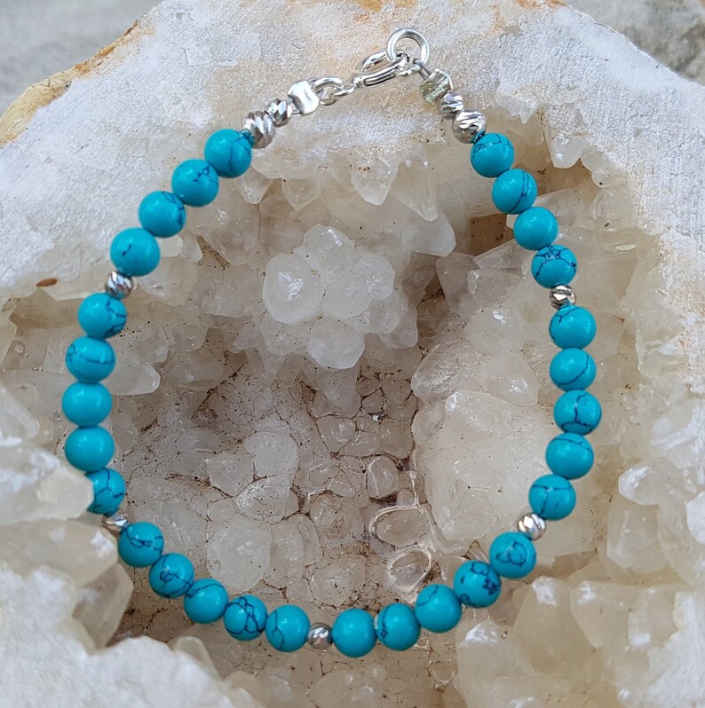 Puede incluir: Un brazalete de piedras turquesa con cuentas de plata y un cierre de plata. El brazalete est&aacute; sobre un fondo blanco.