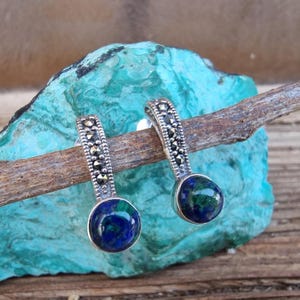 Puede incluir: Un par de pendientes de plata con diseño de marcasita y gotas redondas de piedras preciosas azul-verdes. Los pendientes se exhiben en una rama de madera sobre un fondo turquesa.