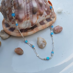 May include: A silver chain necklace with blue opal beads. The necklace is layered and has a delicate design. The necklace is photographed on a white surface with a seashell and small stones.
