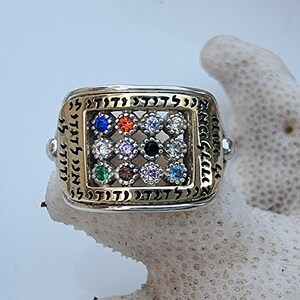 May include: A silver ring with a gold-toned square inset featuring a grid of colorful gemstones. The ring is inscribed with Hebrew characters.