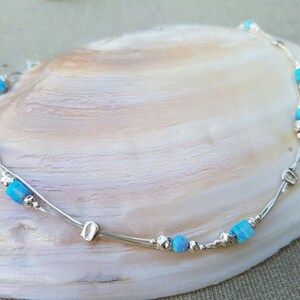 May include: A delicate silver anklet with two strands and small blue opal beads. The anklet has a silver charm in the shape of a square.