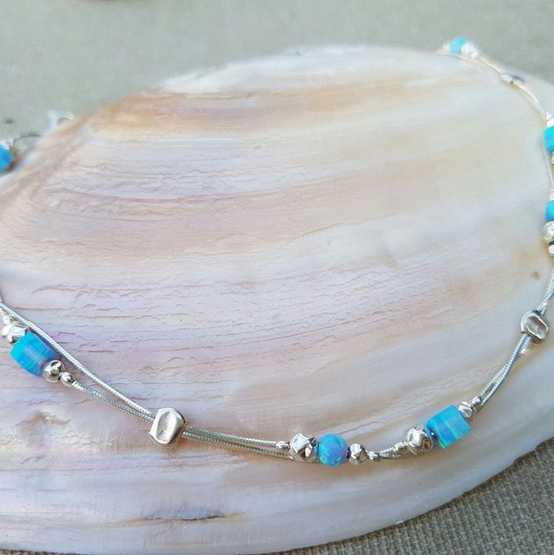 May include: A delicate silver anklet with two strands and small blue opal beads. The anklet has a silver charm in the shape of a square.