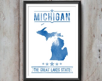 May include: Framed print featuring a blue watercolor map of Michigan, with the state name above and "The Great Lakes State" below. The print is in a black frame and set against a rustic, light-colored wooden background.