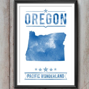May include: Framed print featuring a blue watercolor map of Oregon, with the state name above and "PACIFIC WONDERLAND" below. The artwork is set against a white background, framed in black, and mounted on a wooden wall.