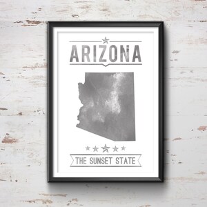 May include: Framed print featuring the state of Arizona in gray, with the state name and "The Sunset State" text. The artwork is in a black frame and is displayed against a white, weathered wood background.