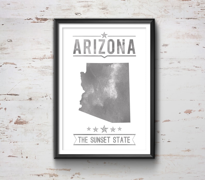 May include: Framed print featuring the state of Arizona in gray, with the state name and "The Sunset State" text. The artwork is in a black frame and is displayed against a white, weathered wood background.