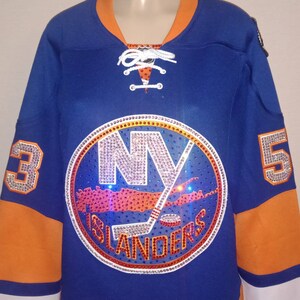 May include: A blue hockey jersey with the New York Islanders logo in rhinestones. The logo is a circle with the letters "NY" in the center and the words "Islanders" below. The jersey has orange and white trim.