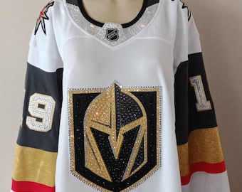 Puede incluir: Jersey de hockey blanco con mangas negras y doradas. El jersey presenta un gran logotipo de un casco de caballero con una 'V' en su interior, incrustado con diamantes de imitación. El número '91' está en las mangas. El logotipo de la NHL está en el pecho.