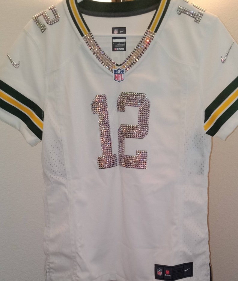 Puede incluir: Camiseta de f&uacute;tbol blanca con ribetes verdes y dorados. El n&uacute;mero 12 est&aacute; en la parte delantera de la camiseta y est&aacute; hecho de pedrer&iacute;a. El logotipo de la NFL est&aacute; en el pecho de la camiseta.