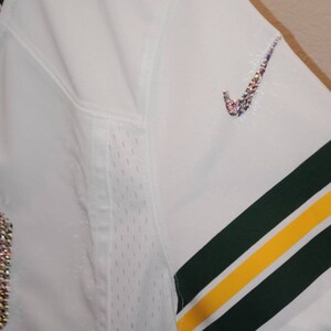 Puede incluir: Camiseta de f&uacute;tbol blanca con rayas verdes y amarillas en las mangas. El n&uacute;mero "1" est&aacute; en la parte delantera de la camiseta y est&aacute; decorado con diamantes de imitaci&oacute;n. El logotipo de Nike est&aacute; en el hombro derecho y tambi&eacute;n est&aacute; decorado con diamantes de imitaci&oacute;n.