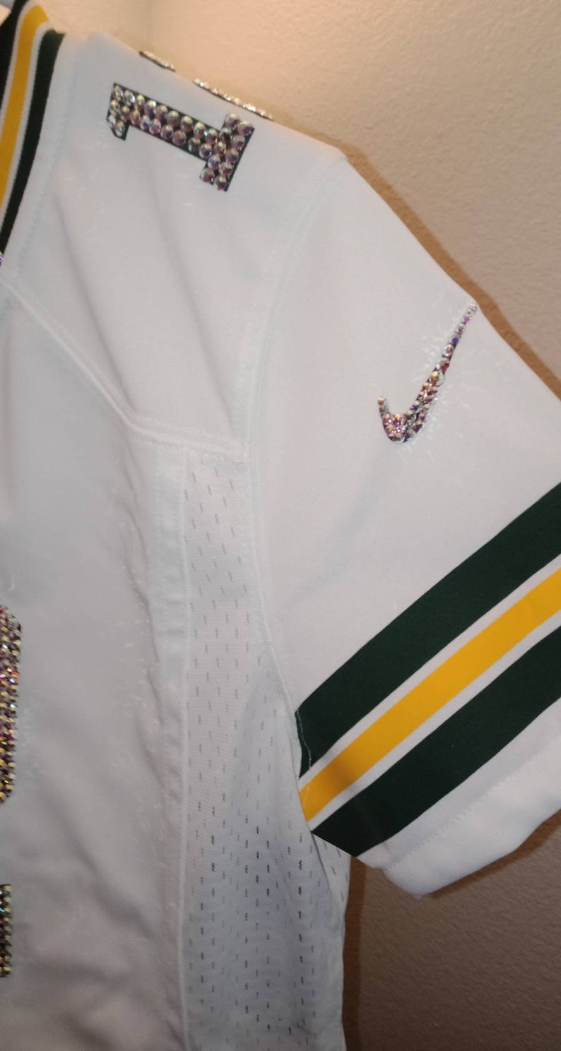 Puede incluir: Camiseta de f&uacute;tbol blanca con rayas verdes y amarillas en las mangas. El n&uacute;mero "1" est&aacute; en la parte delantera de la camiseta y est&aacute; decorado con diamantes de imitaci&oacute;n. El logotipo de Nike est&aacute; en el hombro derecho y tambi&eacute;n est&aacute; decorado con diamantes de imitaci&oacute;n.