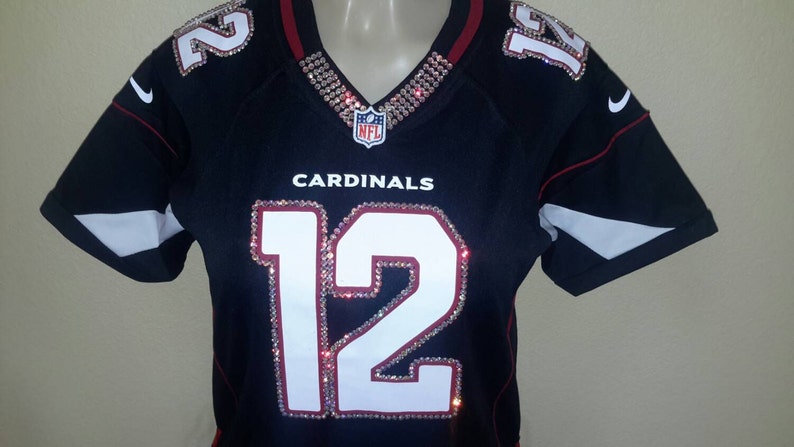 May include: A black football jersey with red trim and white lettering. The jersey has the number "12" on the front, the word "CARDINALS" above the number, and the NFL logo above the word. The jersey is decorated with rhinestones.