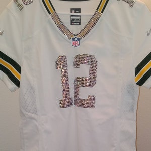 Puede incluir: Camiseta de f&uacute;tbol blanca con ribetes verdes y amarillos. El n&uacute;mero 12 est&aacute; en la parte delantera de la camiseta y est&aacute; hecho de pedrer&iacute;a. El logotipo de la NFL est&aacute; en la camiseta.