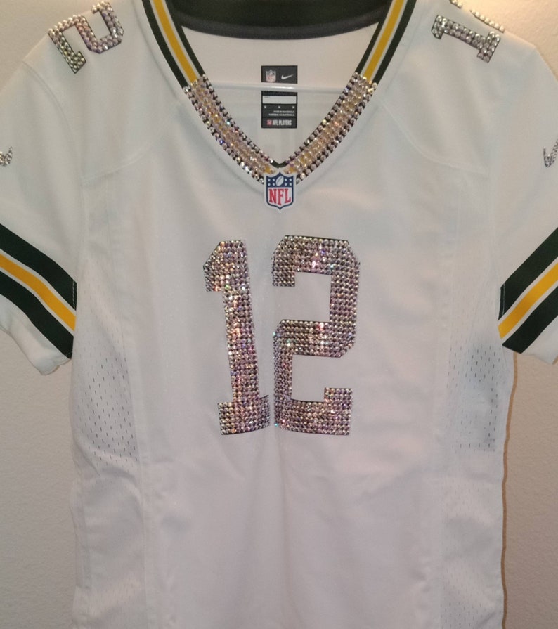 Puede incluir: Camiseta de f&uacute;tbol blanca con ribetes verdes y amarillos. El n&uacute;mero 12 est&aacute; en la parte delantera de la camiseta y est&aacute; hecho de pedrer&iacute;a. El logotipo de la NFL est&aacute; en la camiseta.