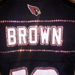 May include: A black jersey with a red cardinal logo and the name "BROWN" spelled out in rhinestones. The jersey has white accents on the sleeves.