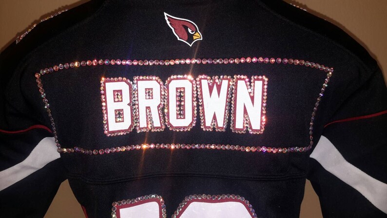 May include: A black jersey with a red cardinal logo and the name "BROWN" spelled out in rhinestones. The jersey has white accents on the sleeves.