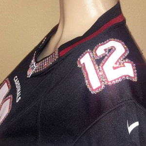 May include: Black football jersey with red trim and a white number "12" with rhinestones on the shoulder. The jersey has the word "Cardinals" on the front.
