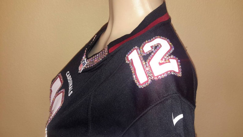 May include: Black football jersey with red trim and a white number "12" with rhinestones on the shoulder. The jersey has the word "Cardinals" on the front.