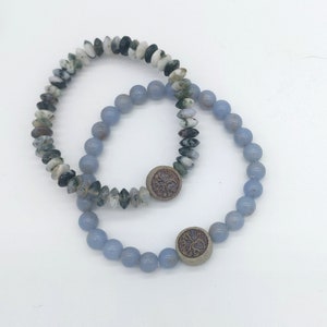 Könnte beinhalten: Zwei Armbänder, eines mit blauen Achatperlen und eines mit grünen und weißen Achatperlen. Beide Armbänder haben einen Metallanhänger mit einem Baummotiv.