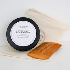 May include: A black jar of beard balm with the text "TUNEITI NATURAL" and "BEARD BALM PATCHOULI" on the label. The jar is sitting on a white surface next to a wooden comb with the text "TUNEITI NATURAL" on it.