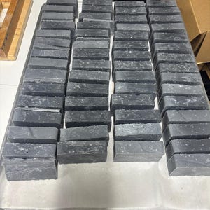 May include: A large collection of rectangular charcoal-colored soap bars arranged in neat rows on a metal tray. Each bar features a solid dark gray base with subtle white speckles distributed across the surface, creating a uniform appearance.